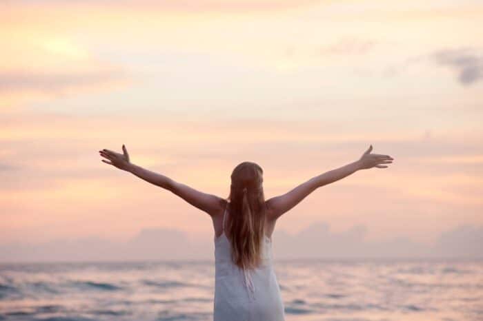 Woman with long blonde hair facing away from the camera, standing in front of the ocean at sunset with her arms open.